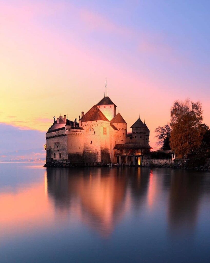 View of Château de Chillon, illuminated at sunset, overlooking the still waters of Lake Geneva.
