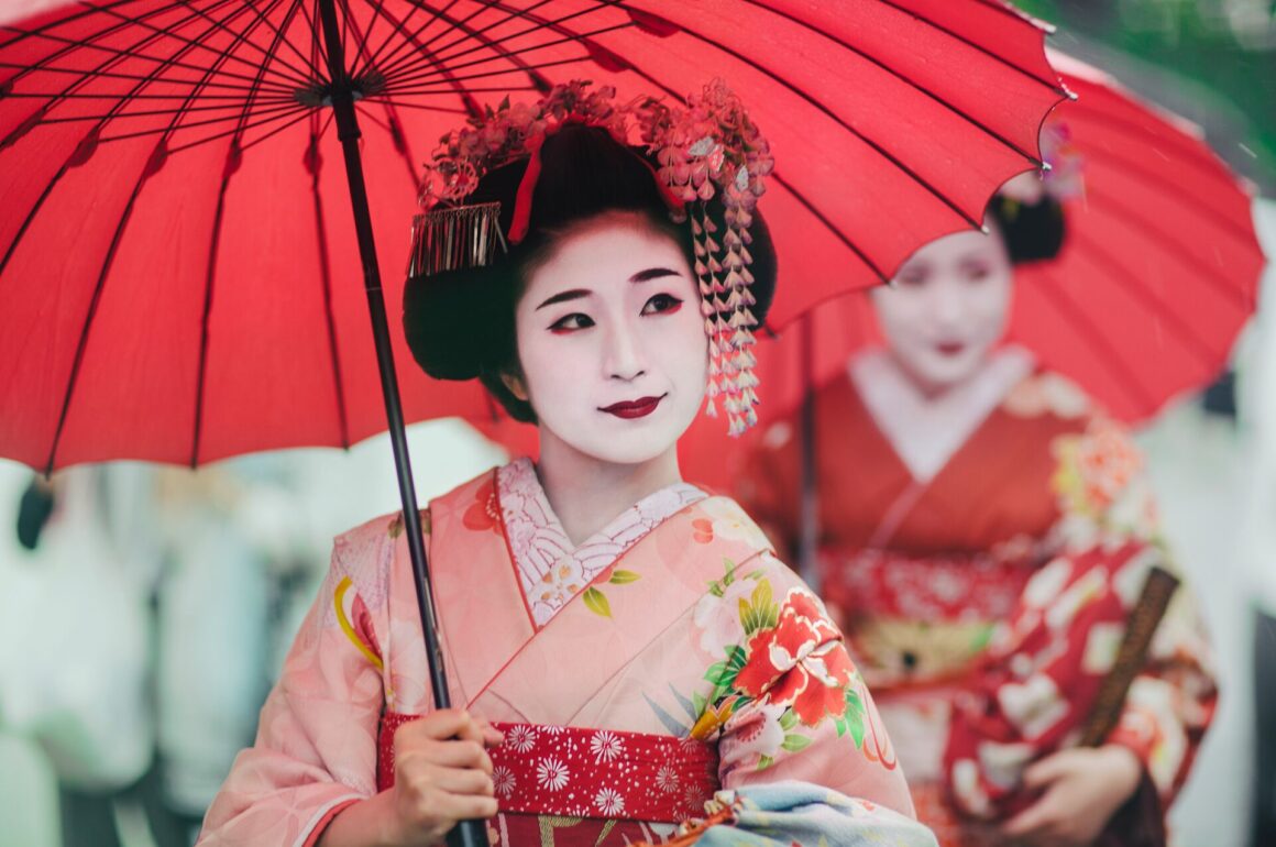 Image of Japanese Geisha wearing kimonos, with traditional make up and red parasols, walking together