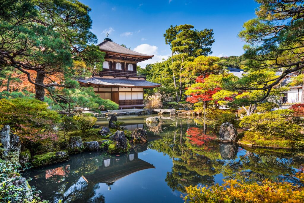 Image of an ornate palace in Kyoto, surrounded by lush, brightly coloured traditional Japanese gardens, with a bright blue pond in front.