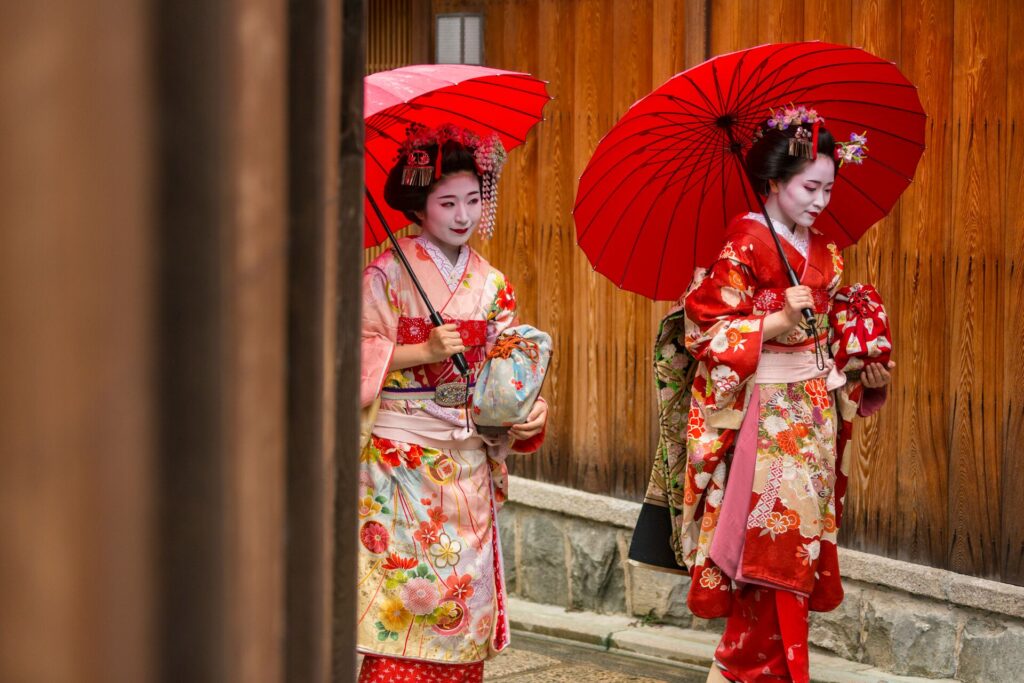 Picture of two Maiko Apprentice Geisha Japanese Women In Traditional Kimonos, waking down the street with red parasols