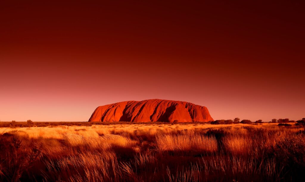 Image of Uluru at sunset, glowing red with the Australian bush in the foreground