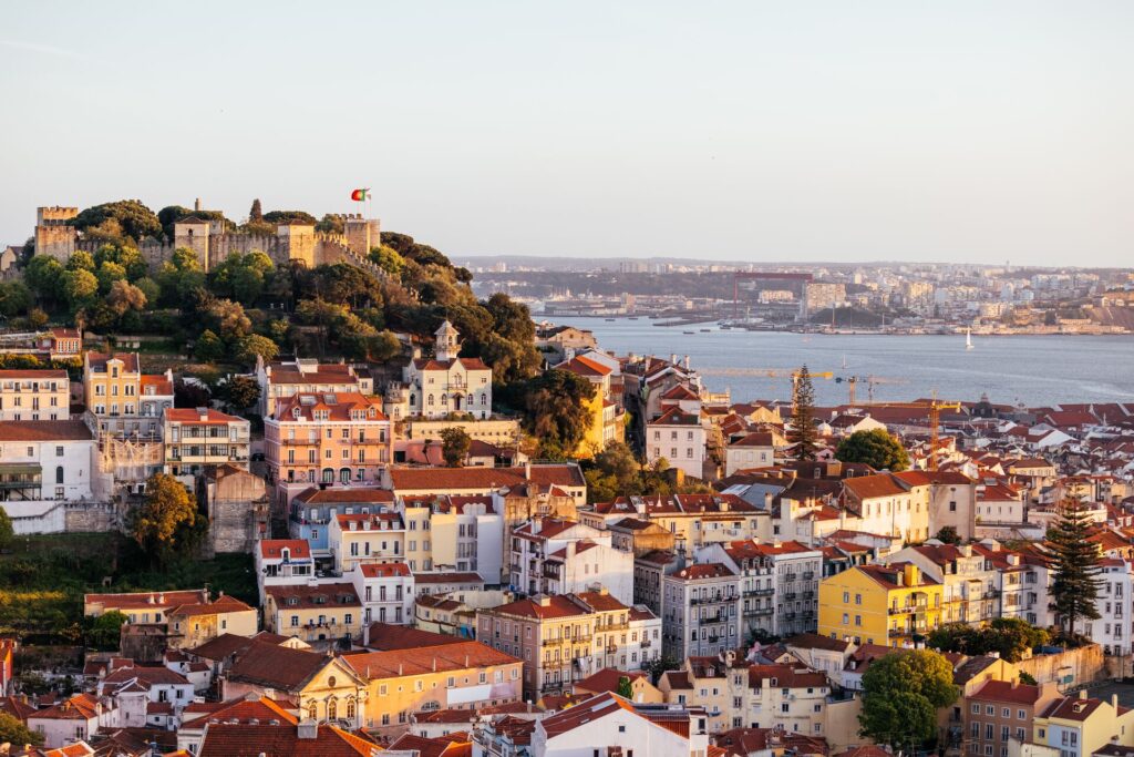 Lisbon cityscape with St George Castle at sunset, showing red roofed houses and water in the background. 