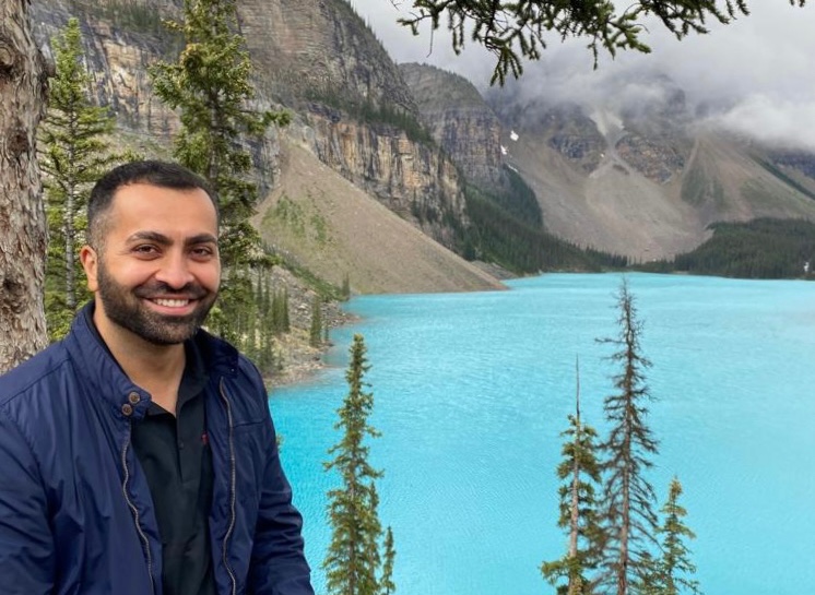 Eastern Canada Travel Concierge Arsen sits in front of an azure lake with grey mountains in the background.