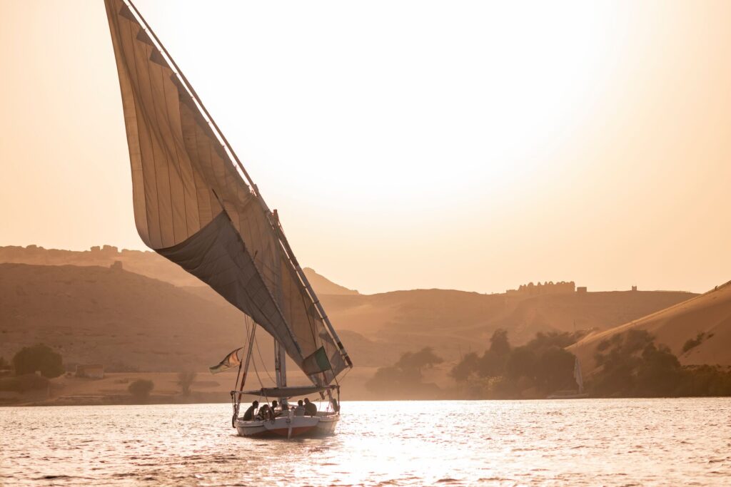 A Felucca boat sails on the nile, with golden tones across the water and landscape, from the sun