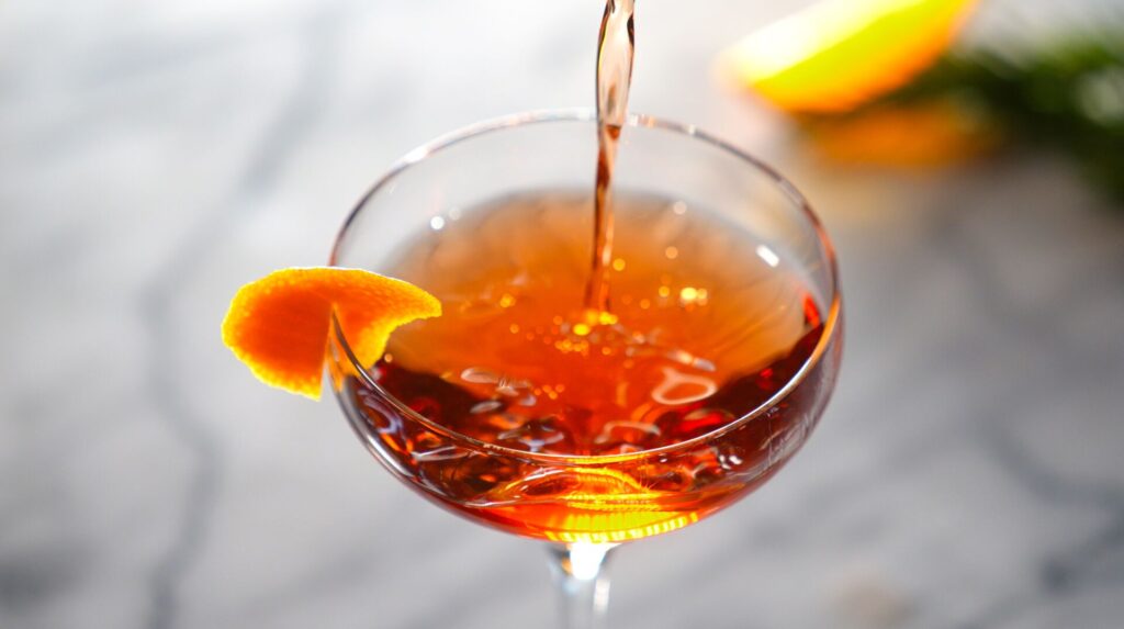 Close up of Negroni cocktail in a tall cocktail glass with a round top, showing bright orange liquid and a twist of citrus rune, against a grey blurred backdrop