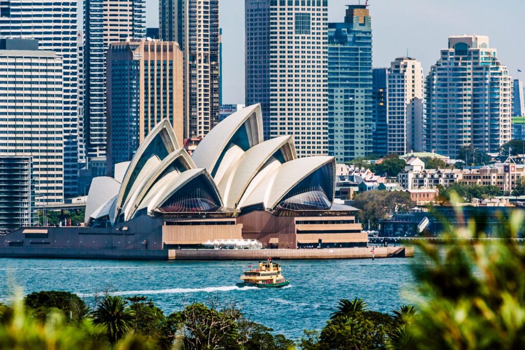 Sydney Opera House shown with the tall CBD buildings behind and the blue harbour waters in front, with greenery close to the camera
