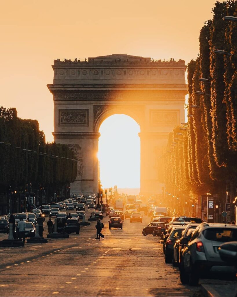 The Arc de Triomphe in Paris with the sun setting behind, creating a warm glow along the Champs-Elysees