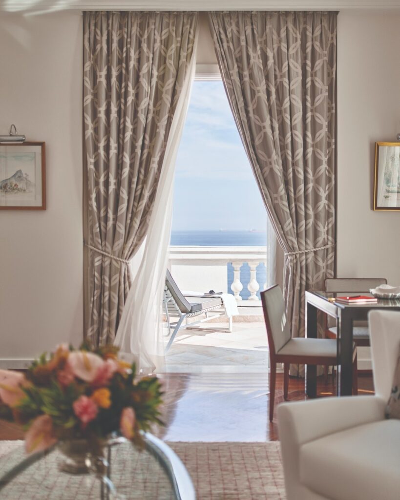 Picture from the inside of a luxury room looking out at the blue ocean through to the terrace, with white and beige furnishings and a bright bunch of flowers in the foreground.
