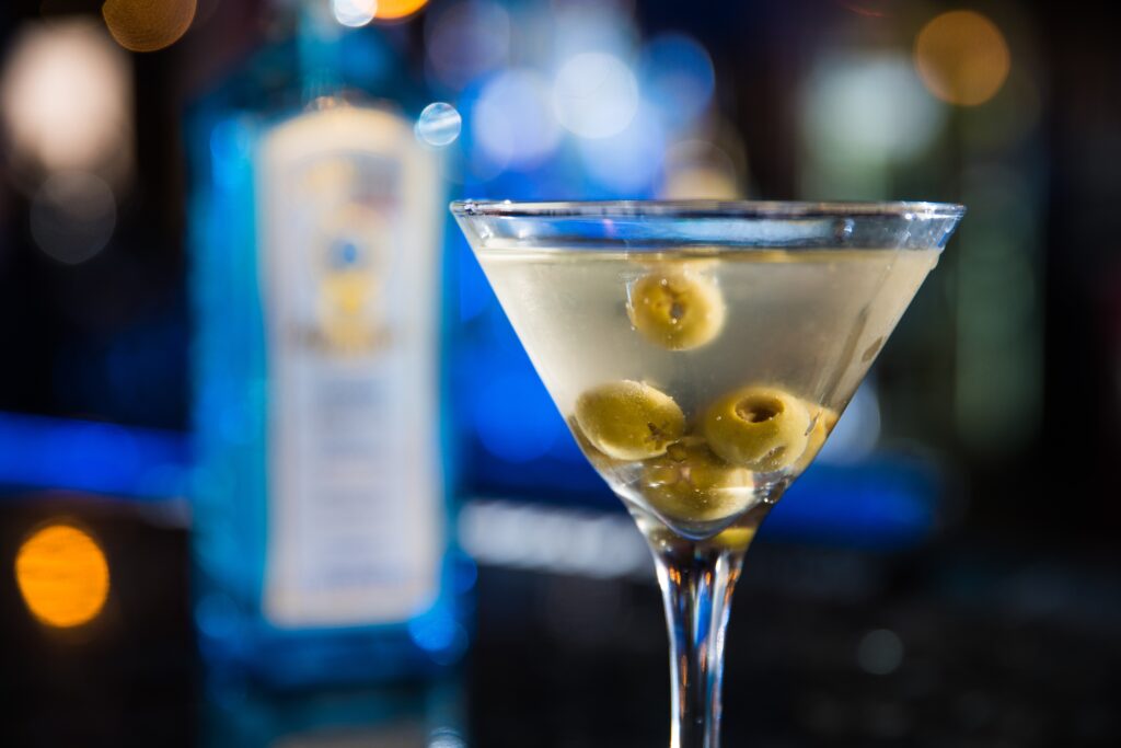 Close up of Martini cocktail in a v shaped tall glass, clear liquid with green olives inside, against a blurred blue background