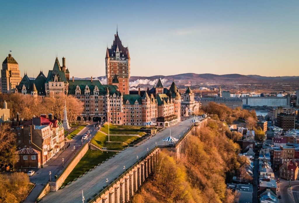 The iconic chateau Le Frontenac hotel in Eastern Canada is pictured perched above the buildings and roads of Quebec, with red brick and green roofs against a dusky blue sky.