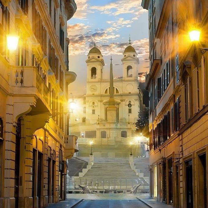 Vis Condotti in Rome, with the Spanish steps rising up to the 16th century church. The sun is rising behind giving a warm glow against a bright blue sky with pink clouds