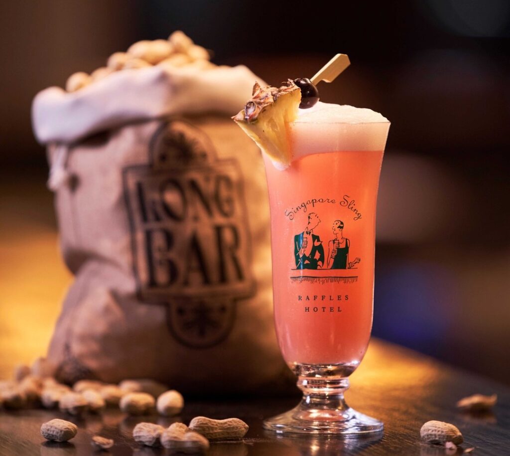 Close up of a bright pink Singapore sling cocktail in a tall glass with a slice of yellow pineapple, in front of an old fashioned peanut bag, both bearing the emblem of the Raffles Hotel.