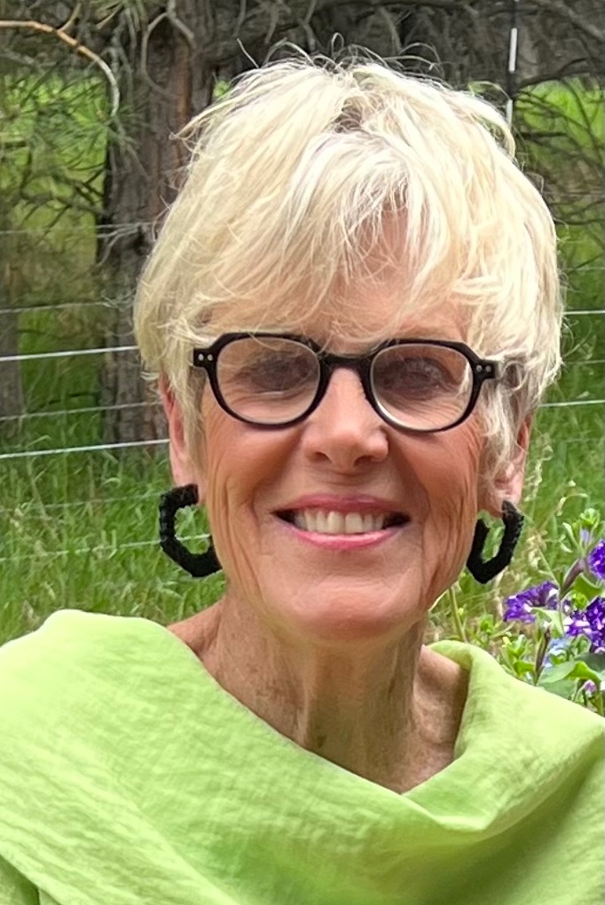 Head shot of Luxury Gold Travel Concierge Ann, wearing a bright green blouse, with short blond hair and dark rimmed glasses and a big smile, with a green field and tree in the background