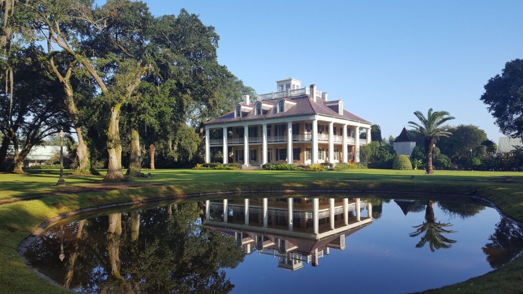 Houmas House in Natchez USA is pictured behind a large blue lake, the houses white columns prominent showing its antebellum style. green trees are on either side. 