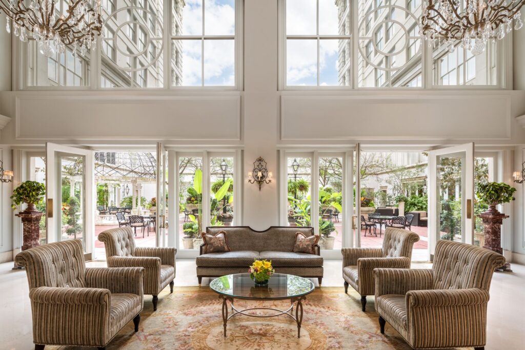 The front room of the Ritz Carlton Hotel, New Orleans is shown with its high windows and white and bright interior color scheme and lush furnishings, with sunlight streaming in