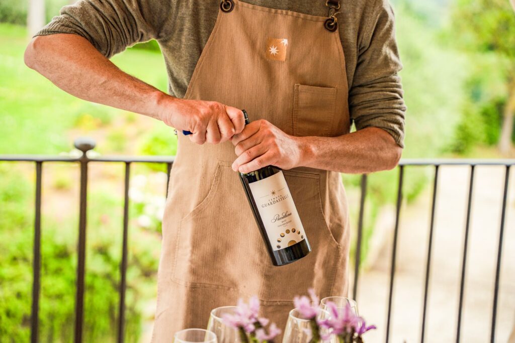 A man opens a bottle of red wine, shown from the next down wearing a brown apron with a blurred green garden behind him. 