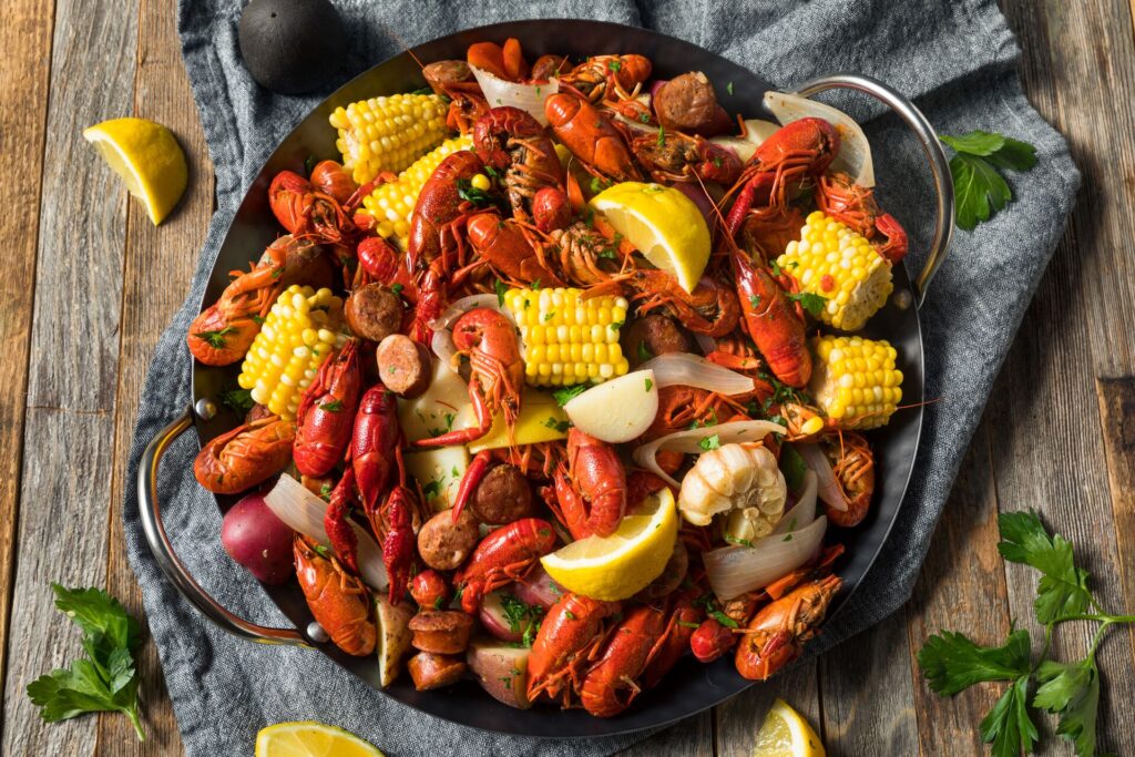 A colourful plate of Crawfish gumbo, with bright red crawfish, yellow sweetcorn, yellow lemons in a big dish on a wooden talbel, with lemons and herbs around the sides