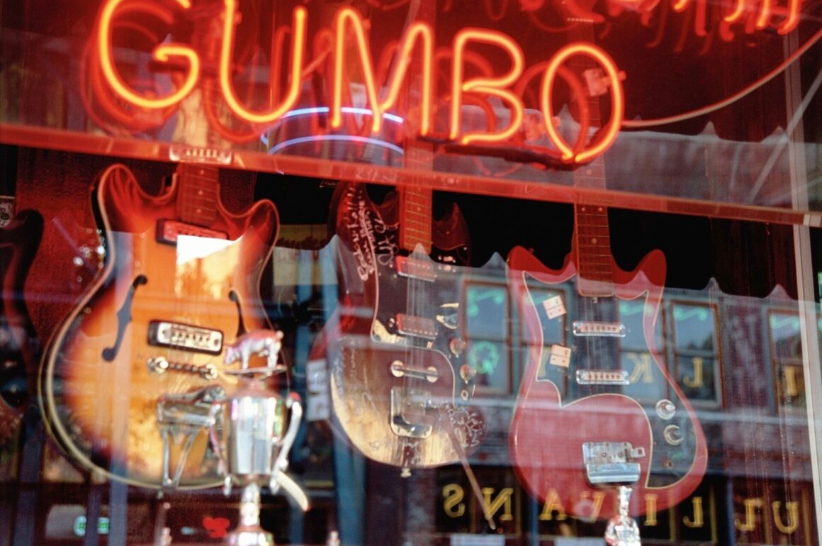 Three guitars are shown in a shop window with a red gumbo sign above them, reflecting the road in a red and orange style picture