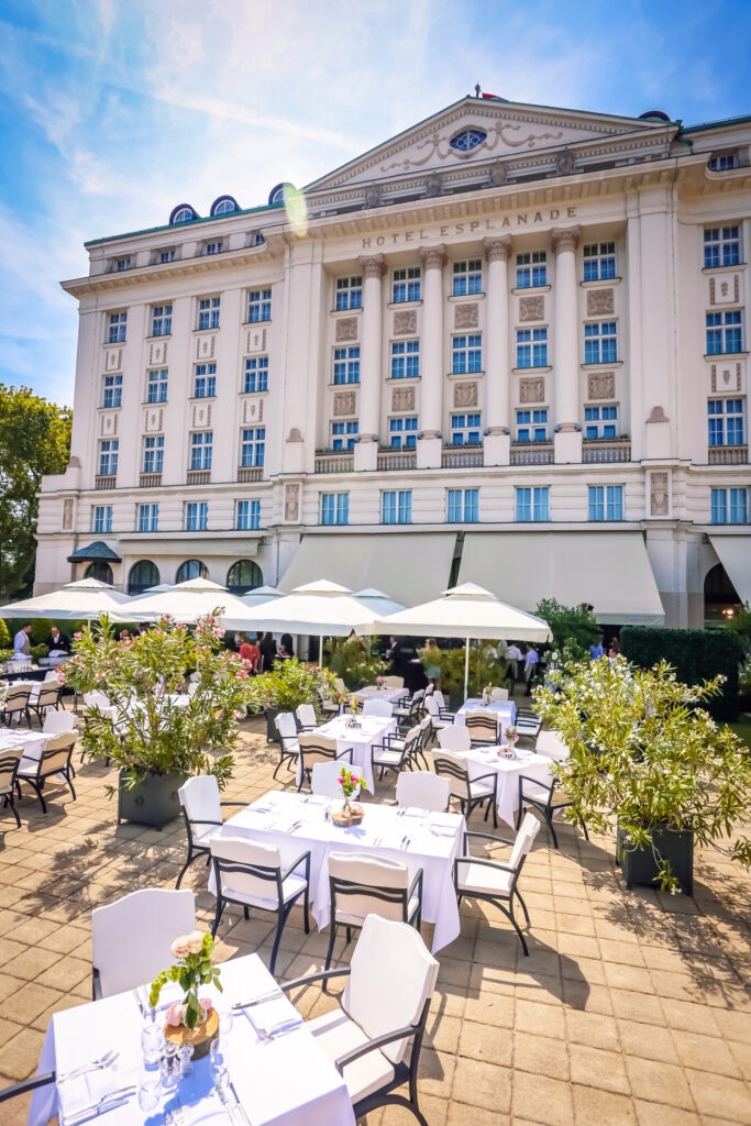 The Oleander terrace of the Hotel Esplanade Zagreb, basking sunshine shows white tables and parasols set against a tiled floor, with the hotel in the background