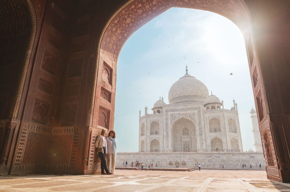 The Taj Mahal is pictured through a large red brick archway with the sun siding brightly.