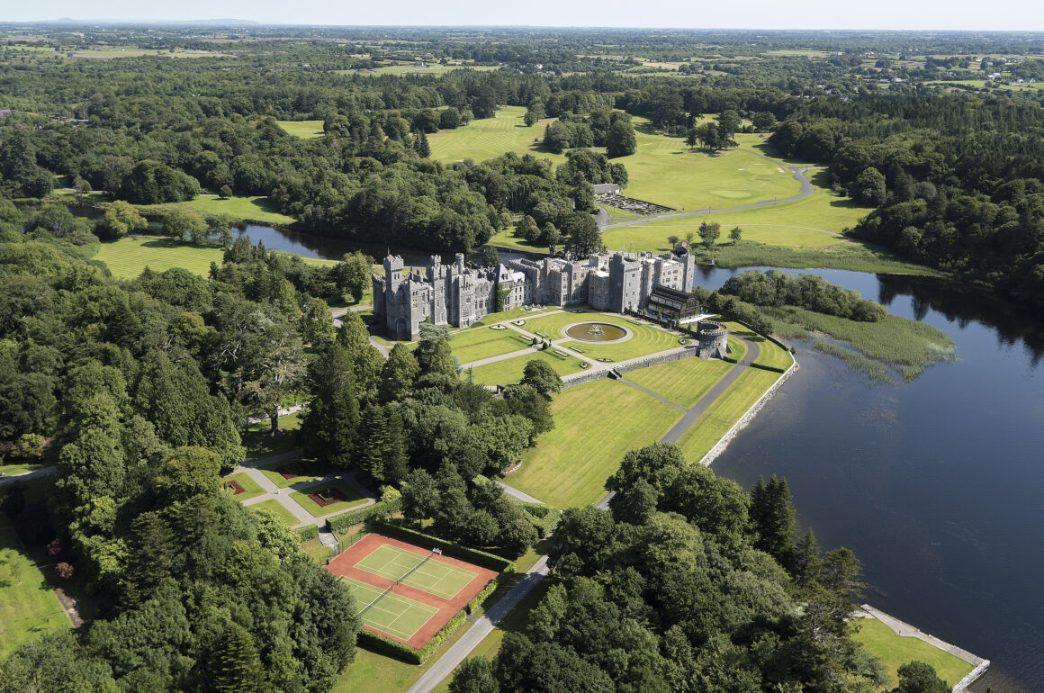 Aerial view of Ashford Castle, showing green gardens, the blue lake, tall trees and tennis courts