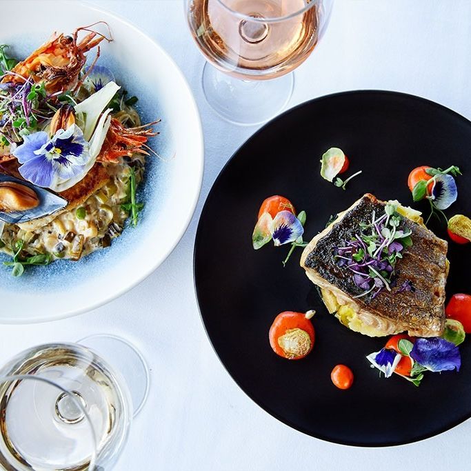 Two elegant seafood dishes shot from above, with fish, bright coloured garnish, flowers petals and wine glasses by the side.
