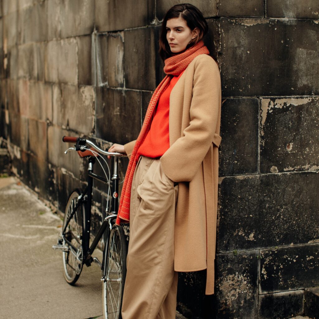 A woman wearing cashmere in warm colours stands by a wall with a bicycle next to her
