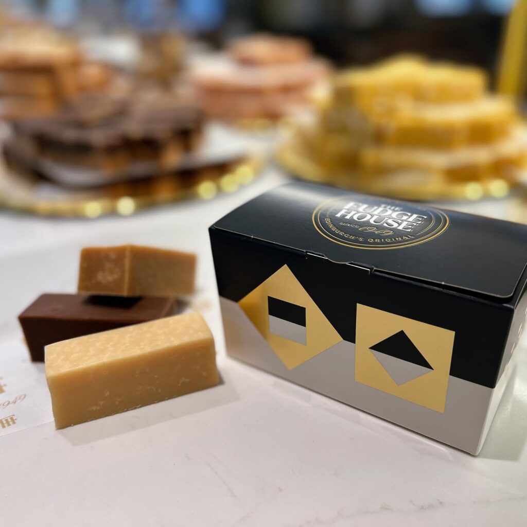 Pieces of brown fudge sit next to a Fudge House presentations box, with blurred candies in the background.