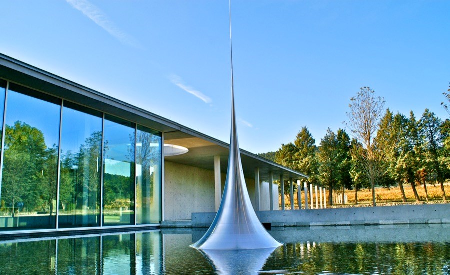 A silver metal spire rises out of a water feature, in front of a futuristic glass building with trees in the background