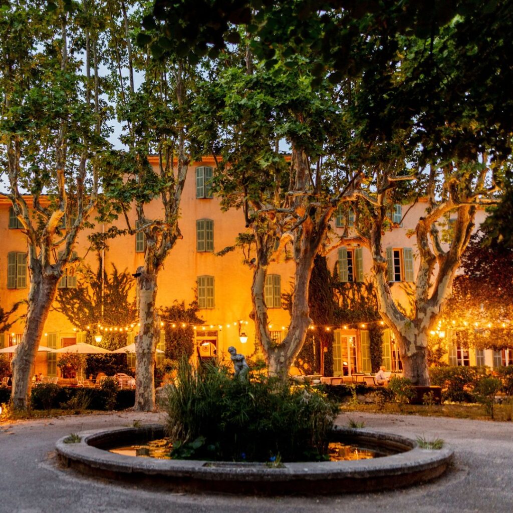 The front of the hotels La Mageleine in Southern France is lit up with warm yellow colours with trees in the foreground and a small ornate pond.