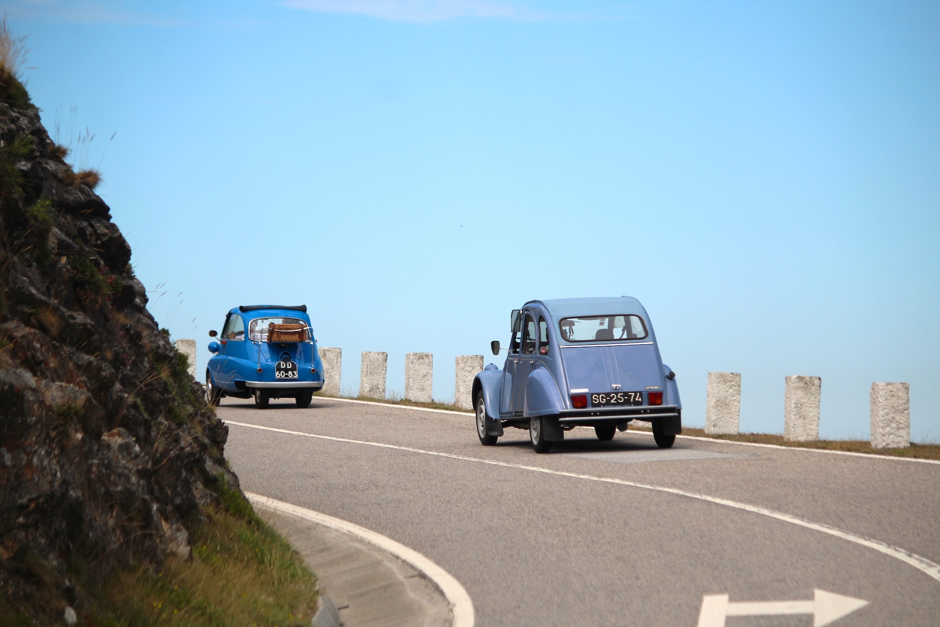 Two classic cars driving up a winding, cliff-side road in France