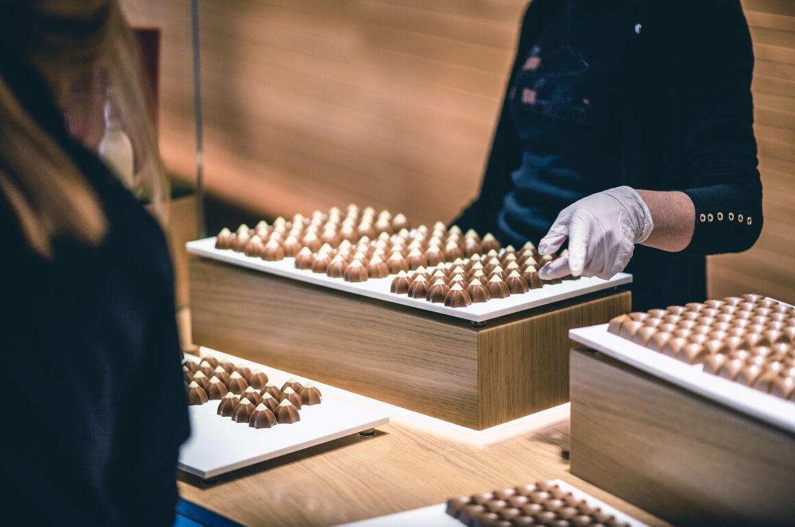 Rows of carefully created chocolates sit in trays, with a white gloved hand showing them off