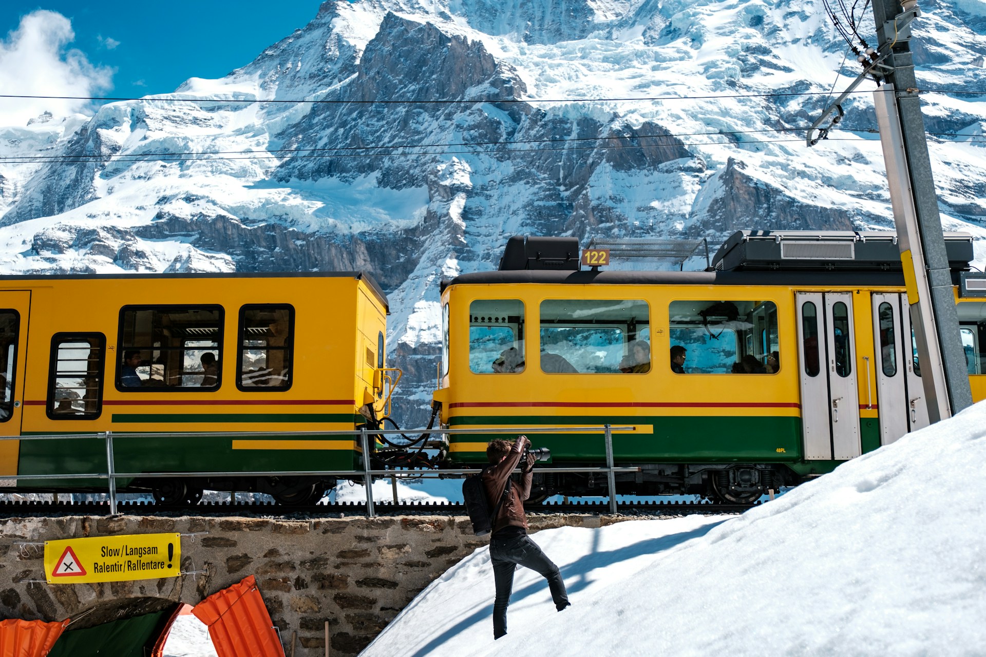 A photographer taking photos of the Jungfrau train. Side view of the train as it passes through the frame, mountains in the background